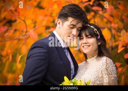 Sposa biraciale e sposo insieme all'aperto dopo il matrimonio autunnale con brillanti foglie arancioni colorate sullo sfondo Foto Stock