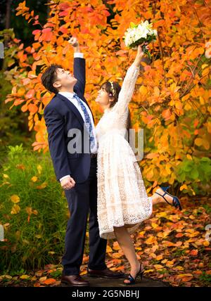 Sposo e sposo biraciale appena sposato sollevando le braccia che si acclamano dopo il matrimonio mentre si levano in piedi all'aperto in autunno, foglie di arancio luminoso in background Foto Stock