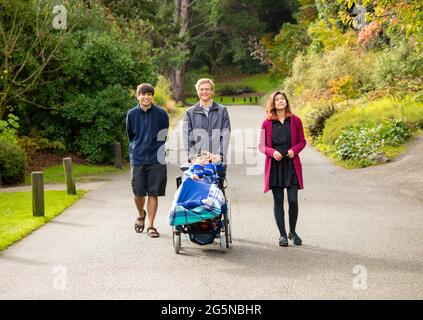 Il padre caucasico cammina con i bambini biraciali attraverso il parco in primavera o in autunno, spingendo il figlio disabili in sedia a rotelle. Famiglia multirazziale. Foto Stock