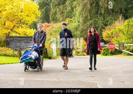 Il padre caucasico cammina con i bambini biraciali attraverso il parco in primavera o in autunno, spingendo il figlio disabili in sedia a rotelle. Famiglia multirazziale. Foto Stock