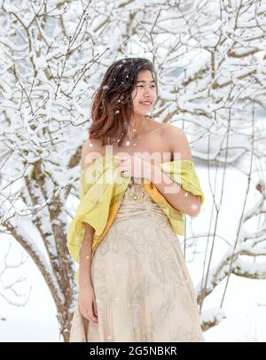 Ragazza adolescente biraciale in abito formale color oro e scialle all'aperto in giornata innevata con rami di alberi ricoperti di neve in background Foto Stock