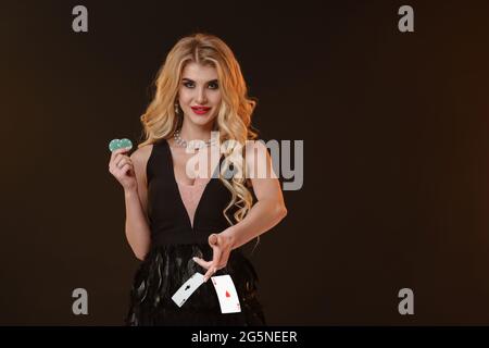 Ragazza bionda in abito nero e collana. Sorridendo, mostrando due fiches verdi, lanciando qualcosa, ponendosi su sfondo marrone. Poker, casinò. Primo piano Foto Stock