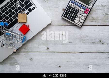 Shopping online o concetto di ecommerce con carrello, borse di carta su computer portatile e calcolatrice su legno bianco con spazio di copia. Foto Stock