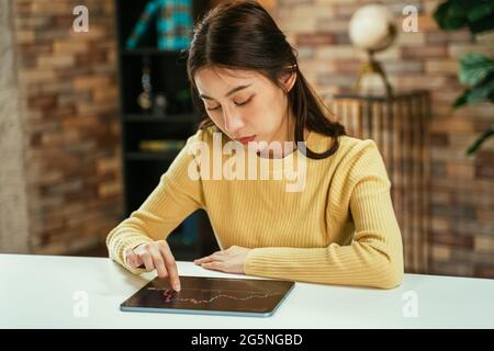 Giovane mediatore asiatico di successo che lavora e legge grafici e dati del mercato azionario utilizzando tablet digitale a casa con concentrazione Foto Stock