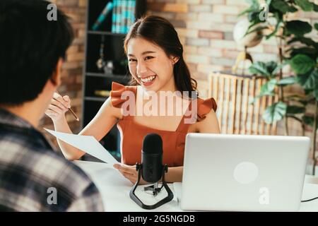 Giovane podcaster asiatico allegro che tiene un questionario mentre intervistando l'ospite maschile al posto di lavoro con microfono e computer portatile Foto Stock