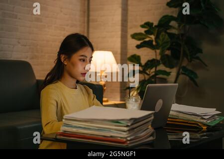 Giovane seria e orientata imprenditrice asiatica femminile lavorando su laptop fino a tarda notte su laptop con file, mentre la fine del progetto per la scadenza a casa Foto Stock