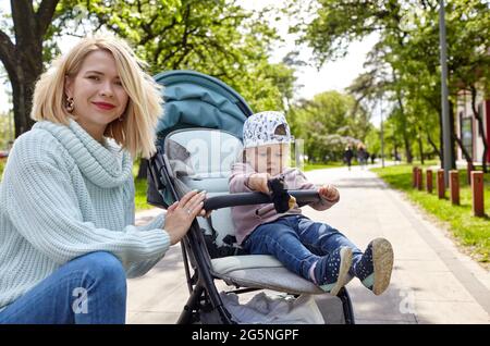 Mamma e figlia sulla natura a piedi presso il parco primaverile. Adorabile bambina in abiti comuni seduta in carrozzina blu. Bambino in buggy Foto Stock