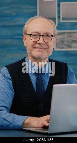 Pensionato manager seduto alla scrivania di fronte alla macchina fotografica sorridente dopo aver digitato al computer portatile lavorando da casa, mentre la moglie anziana leggere un libro in background. Uomo preparato per meeting online utilizzando gadget moden Foto Stock