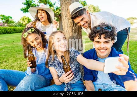 Cinque giovani amici multirazziali che si divertono insieme in un picnic al parco cittadino bevendo birra dalla bottiglia. Giovane donna bionda che prende un auto-ritratto usando Foto Stock