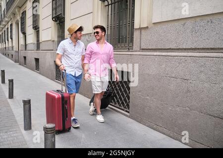 Una coppia gay che porta valigie e va in hotel tenendo le mani in una strada cittadina. Foto Stock