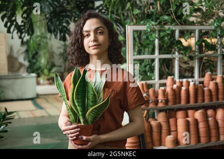 Ritratto di giovane bella donna che tiene in vaso pianta e guardando la macchina fotografica mentre lavora in negozio di fiori Foto Stock