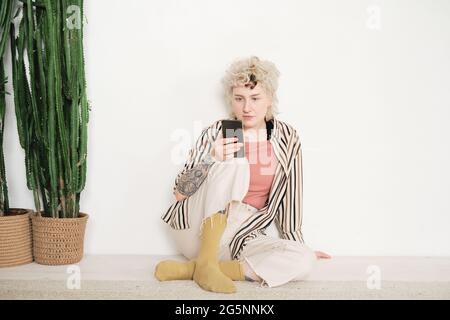 Giovane donna elegante seduta sul pavimento e leggendo online sul telefono cellulare Foto Stock