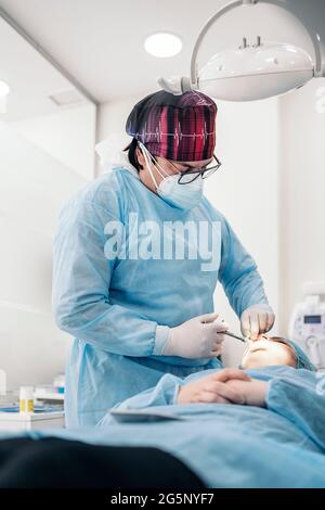 Dentista uomo che indossa maschera facciale e indumenti protettivi che si iniettano pazienti non riconosciuti. Foto Stock