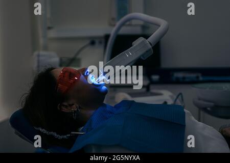 Donna sdraiata su una sedia dentale e indossata occhiali protettivi utilizzando una macchina per sbiancare i denti. Foto Stock