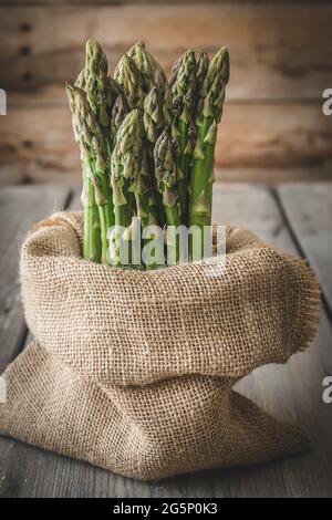 Mazzo di asparagi verdi in un sacco di iuta che si trova su un tavolo di legno, verticale Foto Stock