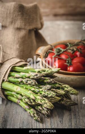 Asparagi verdi su un rustico tavolo di legno, pomodori e un sacco di iuta sullo sfondo, verticale Foto Stock