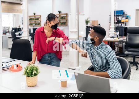 Giovane lavoratore asiatico che parla con il suo collega africano e facendo un gomito a gomito. Indossano maschere facciali a causa del covid19. Foto Stock
