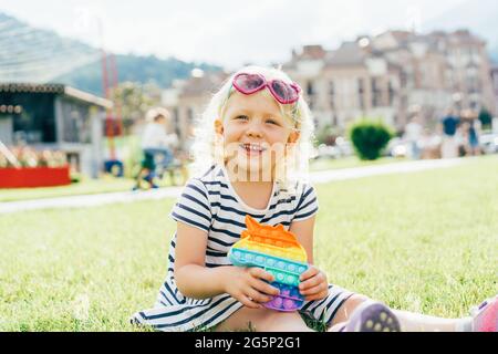 Piccola ragazza felice in occhiali da sole seduto sul prato con anti-stress pop sorride. Il bambino gioca gomma antistress semplice concavità. Foto Stock