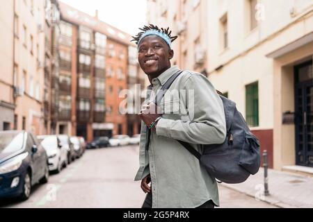 Attraente ragazzo africano con dreadlock e zaino che si indossa camminando in città e guardando davanti. Foto Stock