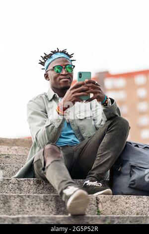 Giovane ed elegante uomo africano seduto nelle scale della città e utilizzando il suo cellulare. Foto Stock