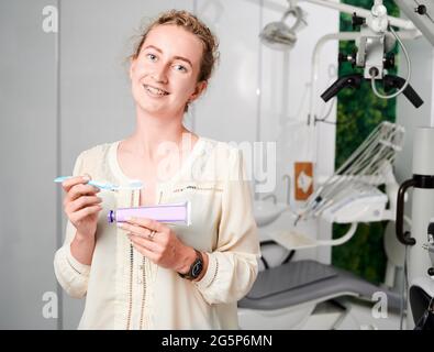 Ritratto di donna in un ufficio dentista ben attrezzato che tiene spazzolino da denti e dentifricio. Giovane ragazza con dentista in ceramica e bretelle metalliche. Spazio di copia. Concetto di stomatologia contemporanea. Foto Stock