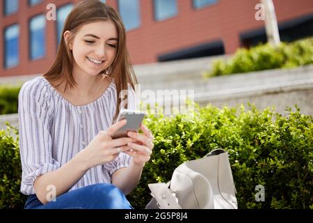 Sorridente giovane donna con telefono cellulare che verifica i social media fuori sede Foto Stock