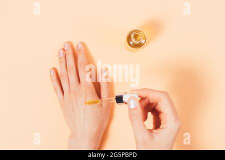 Le mani femminili applicano l'olio cosmetico dalla pipetta alla pelle accanto al flacone aperto, vista dall'alto su sfondo beige. Foto Stock