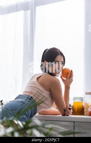 felice giovane donna che tiene arancione e guardando la macchina fotografica in cucina Foto Stock