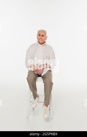 uomo anziano triste in abiti casual seduto su sgabello su bianco Foto Stock