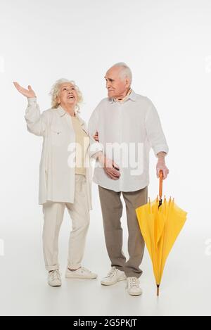 donna anziana stupita con la mano in aria in piedi con il marito che tiene l'ombrello su bianco Foto Stock