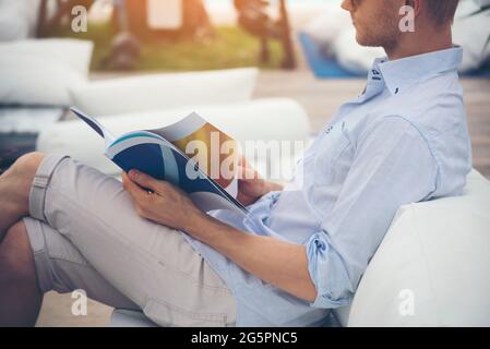 Giovane caucasico che legge libro di rivista sul divano lettore tenendo un libro aperto pagina di carta e la lettura mentre si siede all'aperto sul divano fuori della biblioteca e r Foto Stock