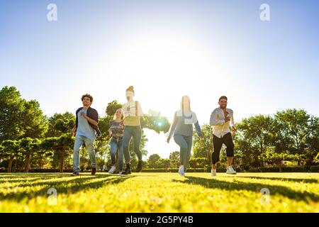 Suggestiva vista ad angolo basso di un gruppo di giovani amici multirazziali di Gen-z che corrono su campo verde con il sole che tramonta alle spalle con effetto luminoso e lungo Foto Stock