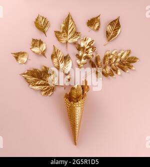 Art. Parete Set botanico. Decorazione stile di vita. Cono gelato in oro con foglie d'oro autunnali, su sfondo rosa. Disposizione piatta. Vista dall'alto. Bella composizione Foto Stock