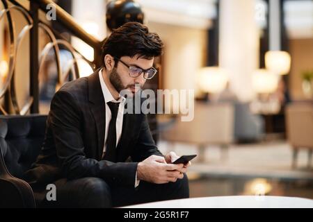 Ritratto di businessman medio-orientale con smartphone mentre si aspetta nella sala dell'hotel, spazio per fotocopie Foto Stock