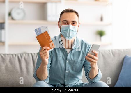 Giovane ragazzo in maschera di prenotazione tour o hotel online, utilizzando smartphone, tenendo in mano il passaporto e biglietti aereo a casa Foto Stock