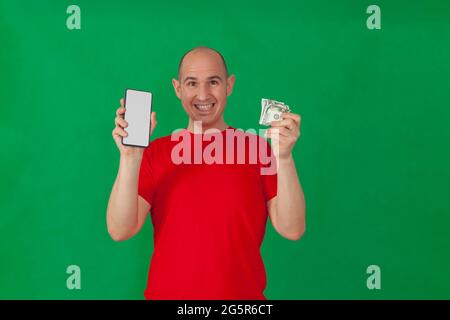 Un calvo caucasico sorridente che indossa una t-shirt rossa che tiene uno smartphone con uno schermo bianco in una mano e 50 dollari di bollette nell'altra. Lo sfondo è Foto Stock