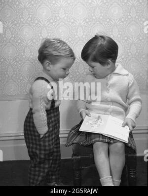 anni '60, storico, che cos'è quel libro che stai leggendo? Un ragazzino in dungree di tartan che mostra interesse nel libro della sorella che ha sulle sue grembe, un libro dell'immagine dei fiori, Inghilterra, Regno Unito. Foto Stock