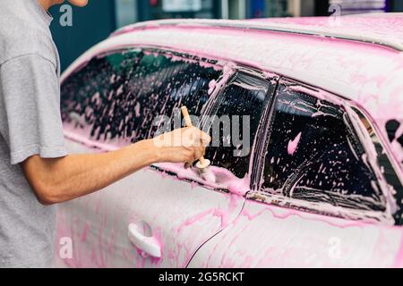 Uomo lavoratore lava i cerchi in lega di un'auto a un autolavaggio, un uomo lava i cerchi dell'auto con schiuma rosa e una spazzola Foto Stock