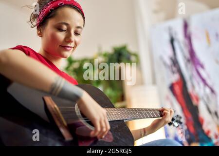 Giovane donna creativa che suona la chitarra acustica mentre si passa il tempo a casa da sola Foto Stock