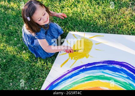 Bambina di 2-4 anni dipinge l'arcobaleno e il sole su un grande foglio di carta seduto su prato verde Foto Stock