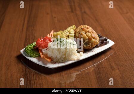Cotoletta di pollo a Kiev impanata in pangrattato, con riso, patatine fritte, pomodori e cetrioli in un piatto bianco sul tavolo Foto Stock