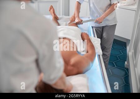 Il terapeuta effettua il massaggio delle gambe con l'attrezzo al paziente dell'uomo con il collega nell'ospedale moderno Foto Stock