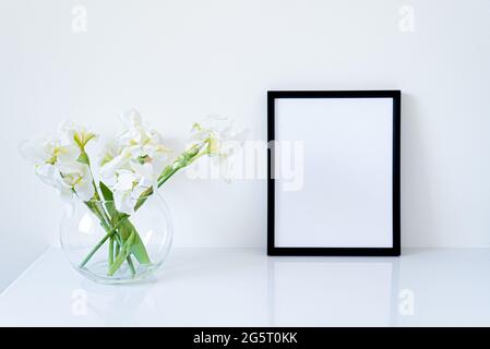 Mockup nero vuoto della cornice. Fiori bianchi iris freschi in vaso di vetro a forma di sfera su tavolo bianco. Sfondo bianco, spazio minimalista ed elegante Foto Stock