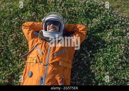 Cosmonauta che indossa tuta spaziale e casco con occhiali da sole che hanno un riposo mentre si trova su erba verde all'aperto Foto Stock