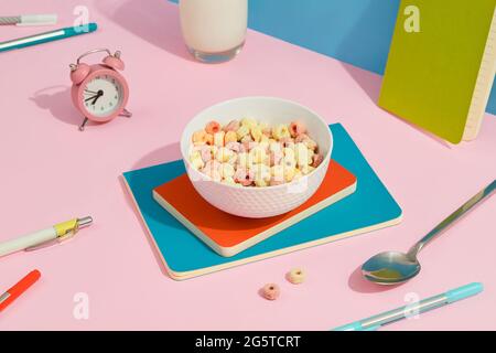 Anelli di grano colorati in una ciotola bianca con cancelleria su tavola rosa. Cibo per la colazione per bambini Foto Stock