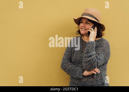 Una donna sorride mentre fa una chiamata sul suo telefono personale, indossa un cappello e si appoggia contro una parete gialla. Immagine urbana Foto Stock