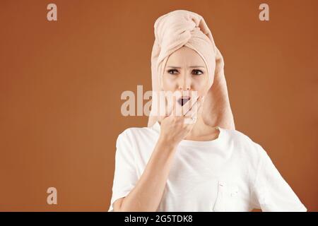 Giovane donna caucasica scioccata con i capelli avvolti in asciugamano da bagno che copre la bocca aperta con la mano su sfondo marrone Foto Stock
