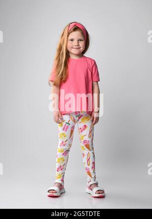 Carino sorridente bambina dai capelli rossi si leva in estate girish indossare t-shirt rosa, pantaloni colorati con stampa e sandali Foto Stock