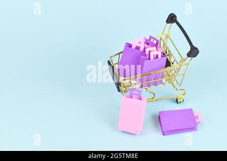 Piccolo carrello d'oro pieno di borse di carta rosa e viola sul lato dello sfondo blu con spazio per la copia Foto Stock