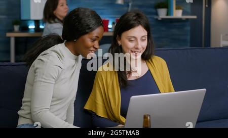 Le donne miste che si divertono a correre trascorrono il tempo insieme guardando il film sul computer portatile mentre si rilassano sul divano. In background donna e uomo bere birra, socializzando a tarda notte durante la festa relax Foto Stock